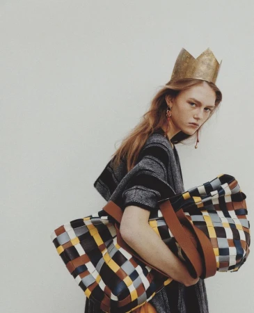 Model wearing a gold paper crown and carrying a large multicolored woven tote bag against an off-white seamless studio backdrop.