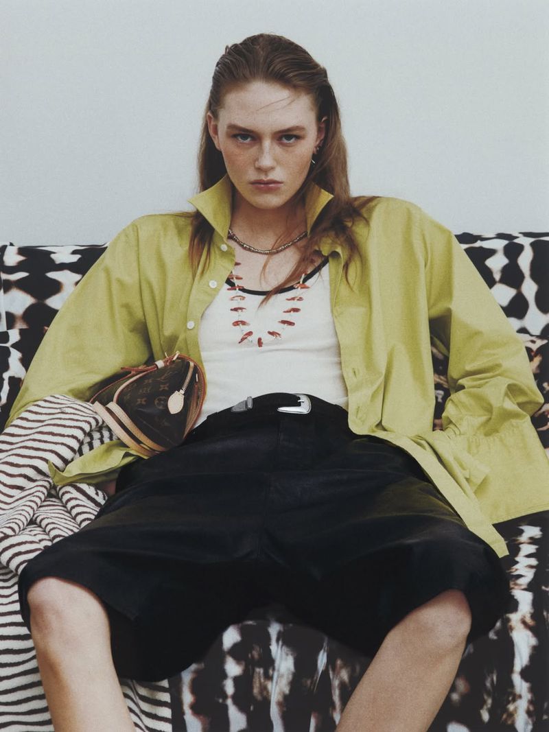 Model Agnes Wahlström seated in an oversized lime green shirt, white tank top, and black shorts for Heroine Magazine.