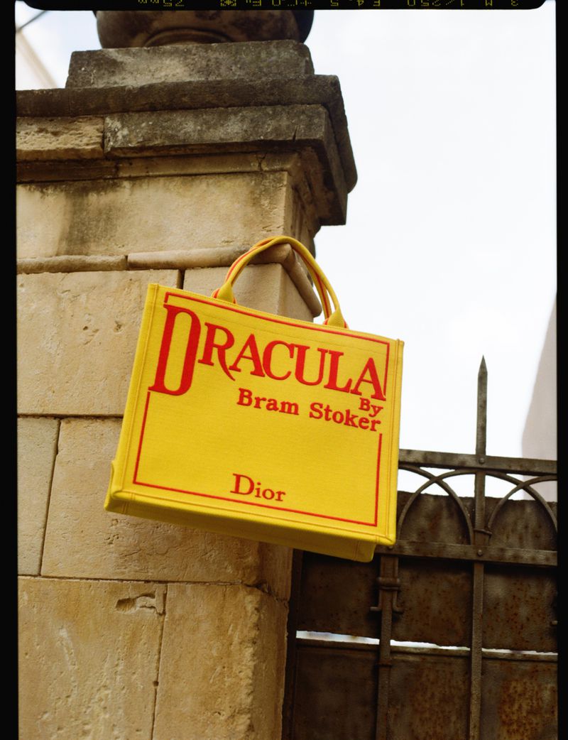 Bright yellow Dior book tote bag with red Dracula typography hanging on a weathered stone pillar beside a rusted iron gate.