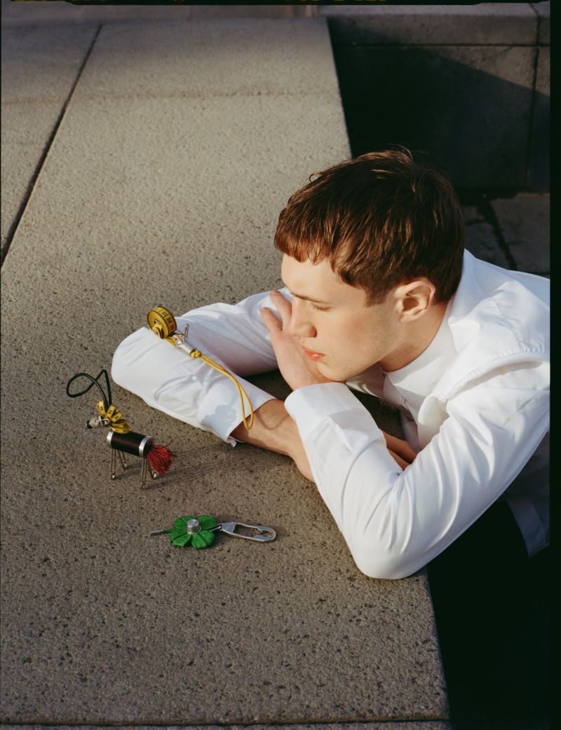 Model Achille Genet in a white shirt resting on a concrete ledge looking at small novelty charms under direct sunlight.