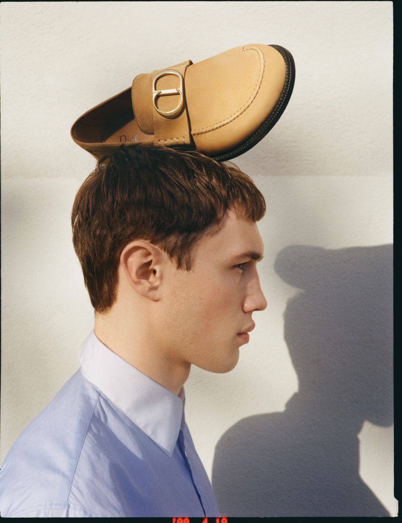 Model Achille Genet in profile wearing a blue collared shirt while balancing a tan leather Dior loafer on his head.