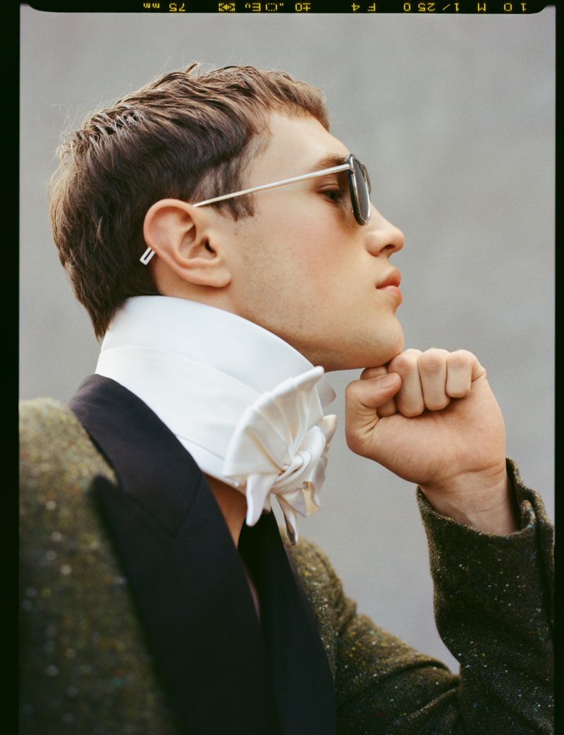 Side profile portrait of a man wearing sunglasses, a high white ruffled collar, and a textured tweed jacket resting his chin on his fist.