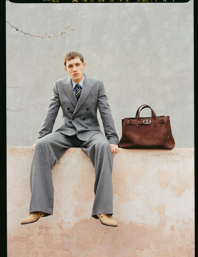 Model Achille Genet wearing a grey double-breasted suit sits on a wall beside a brown suede handbag for Dior Men.