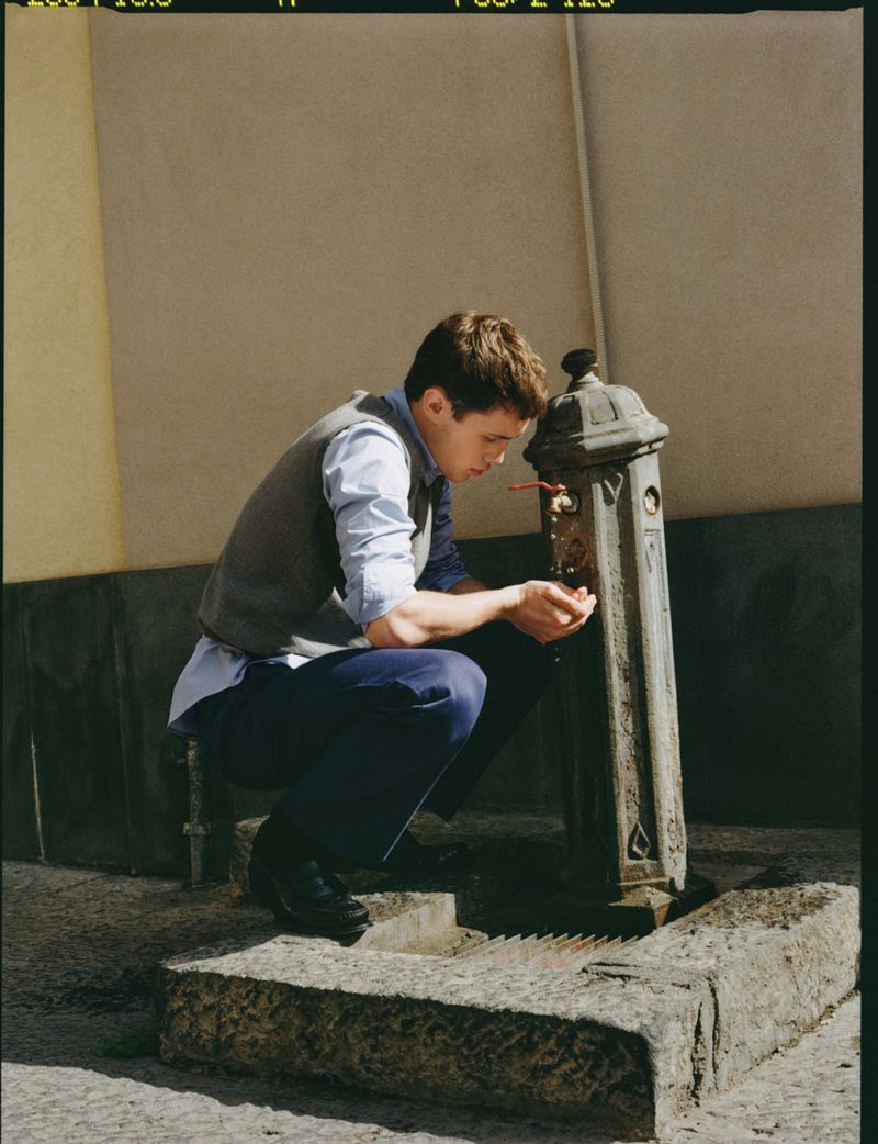 Young man in a grey knit vest and blue trousers crouching to drink from a vintage street water fountain.