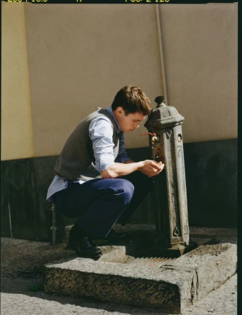 Young man in a grey knit vest and blue trousers crouching to drink from a vintage street water fountain.