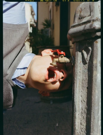 Close-up of a young man drinking directly from an outdoor metal water fountain tap in natural sunlight.