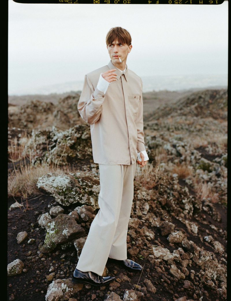 Male model standing on rocky volcanic ground wearing a pale beige collared shirt suit and black shoes with a cigarette.