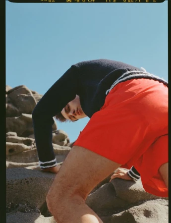 Man bending backward on coastal rock formations wearing bright red shorts and a dark sweater with striped trim.
