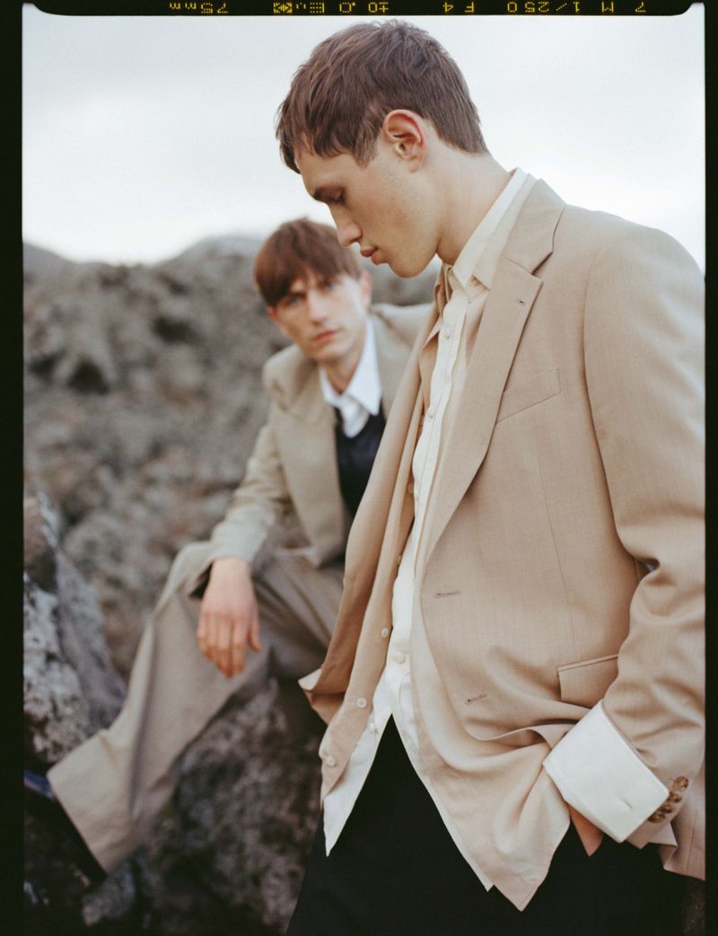 Two male models wearing beige tailored blazers and trousers pose on rocky outdoor terrain under an overcast sky.
