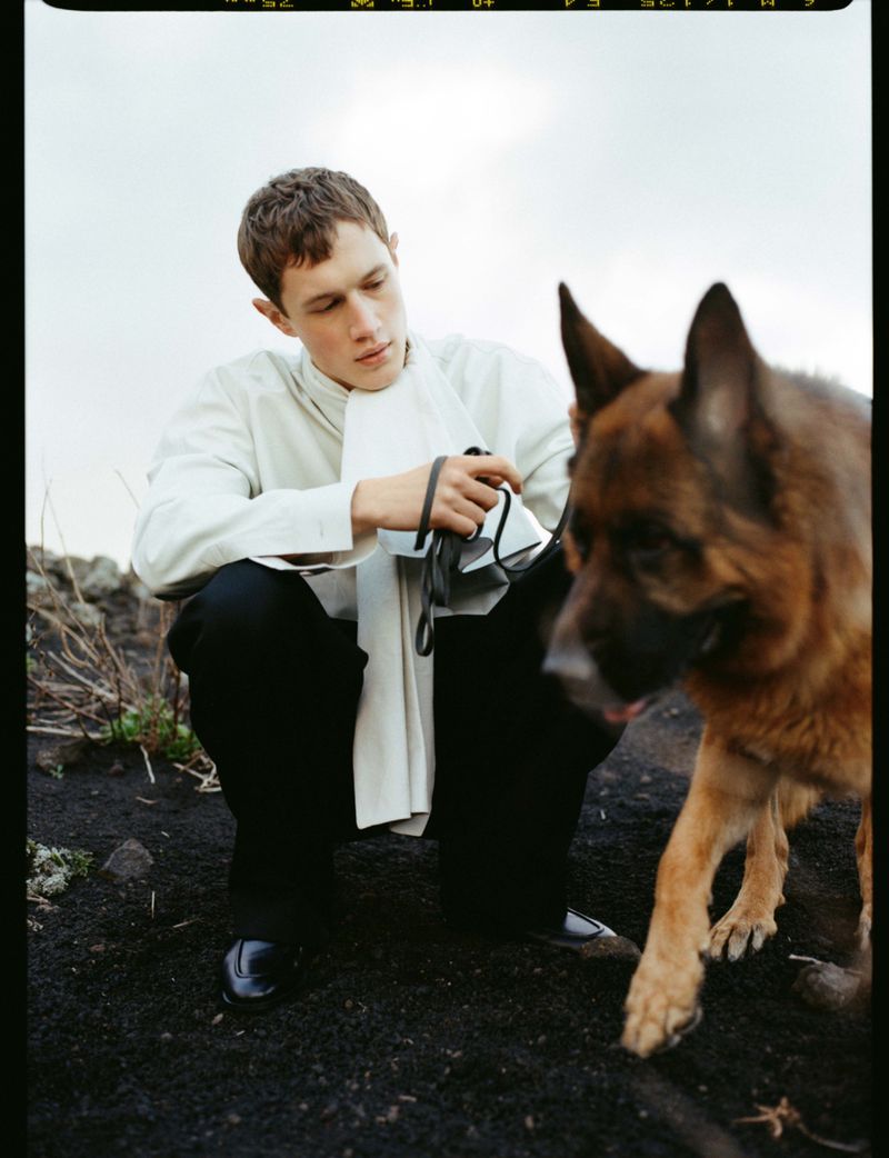 A young man in a white draped shirt and black trousers crouches beside a German Shepherd on dark ground outdoors.