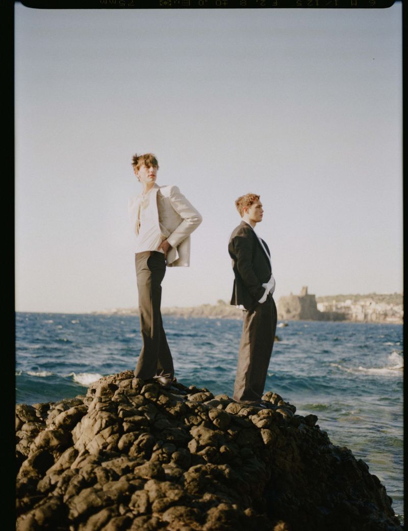 Two male models in tailored suiting pose on a dark volcanic rock formation by the sea under a clear daylight sky.