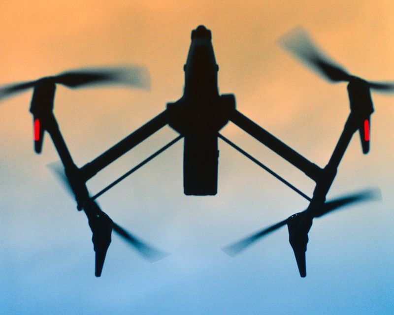 Silhouette of a quadcopter drone with rotating propellers flying against a soft orange and blue twilight gradient sky.
