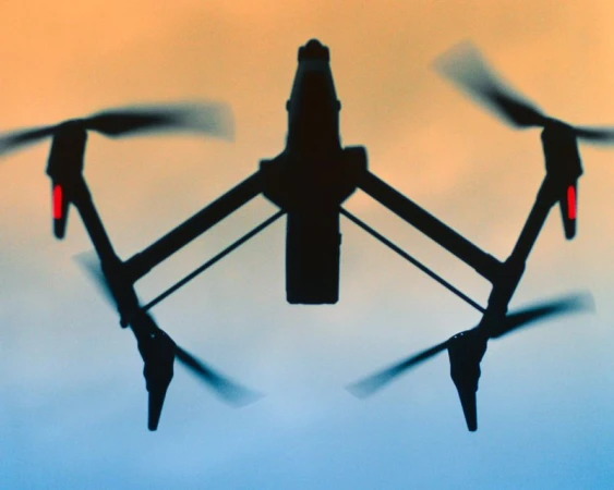 Silhouette of a quadcopter drone with rotating propellers flying against a soft orange and blue twilight gradient sky.