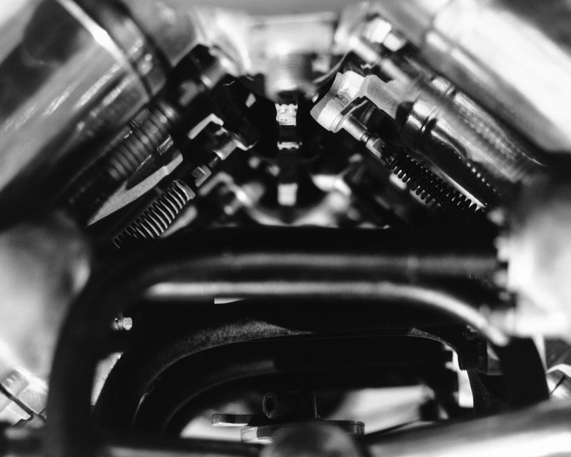 A high-contrast black and white close-up photograph of a mechanical engine with coiled springs, hoses, and polished metallic tubing.