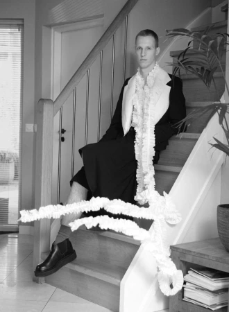 Model Jelle Huizinga in a tailored black suit with long white ruffled scarf seated on a wooden staircase indoors.