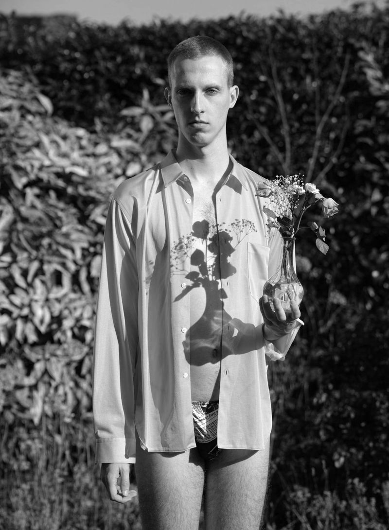 Monochrome portrait of a man in an unbuttoned dress shirt holding a small vase of flowers casting a shadow outdoors.