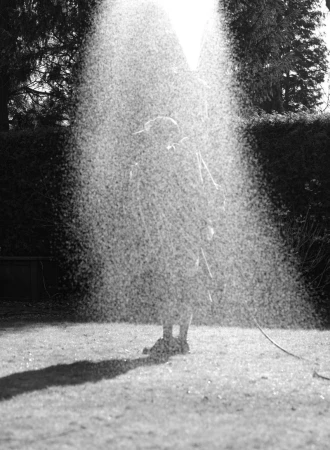 Monochrome photograph of a figure obscured behind a dense spray of water in an outdoor garden setting.