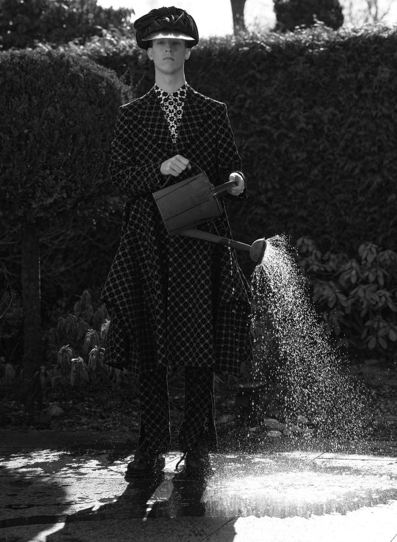 Model standing outdoors wearing a patterned coat and cap, pouring water from a watering can onto the ground