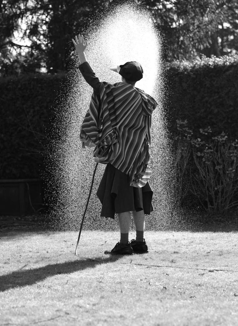 Model viewed from behind in a striped poncho spraying a high arch of water over a lawn in black and white.