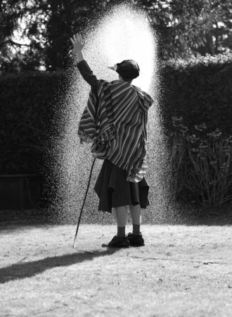 Model viewed from behind in a striped poncho spraying a high arch of water over a lawn in black and white.
