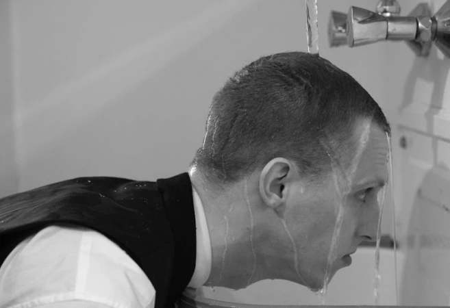 Black and white profile of a man with water pouring over his head from a faucet while wearing a vest and collared shirt.