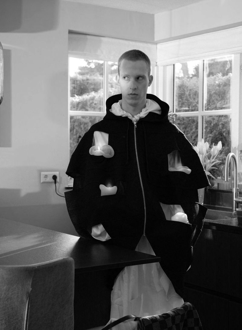 Model Jelle Huizinga wearing an oversized black hooded jacket with glowing candle details in a kitchen setting.