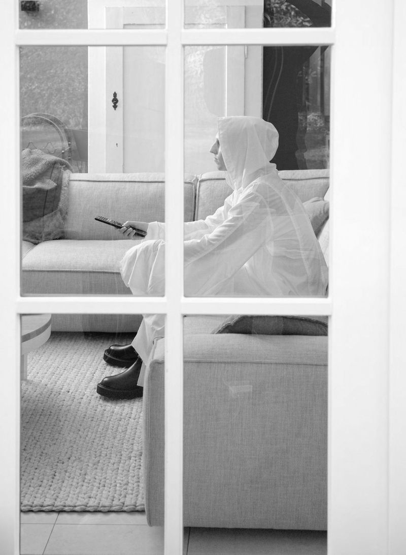 Black and white shot through a window frame of a model in a hooded white garment seated on a living room sofa holding a remote control.