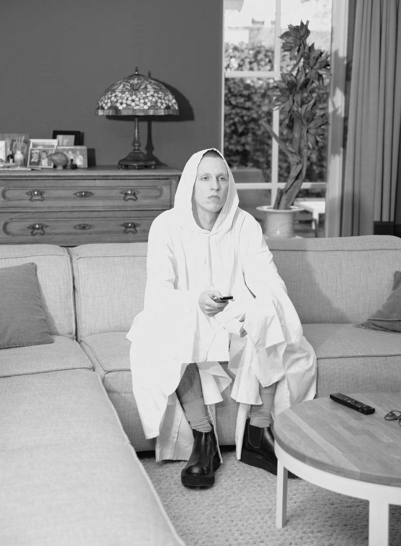 Model Jelle Huizinga seated on a sofa wearing an oversized white hooded garment while holding a remote control in a living room.