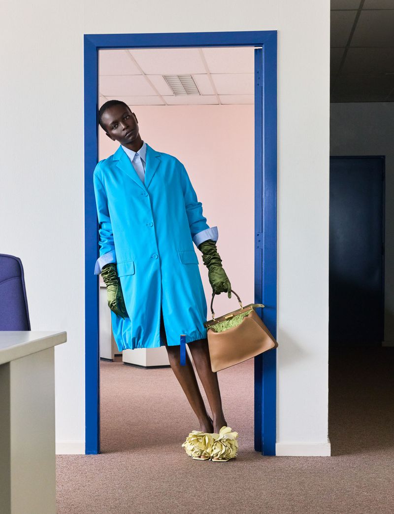 A Black woman in a bright blue coat and long green gloves leans against a blue doorway holding a tan structured handbag.