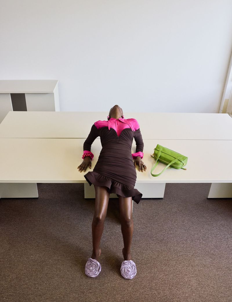 Model leaning back across a white office table wearing a brown and pink dress, floral slippers, and a lime green handbag nearby.
