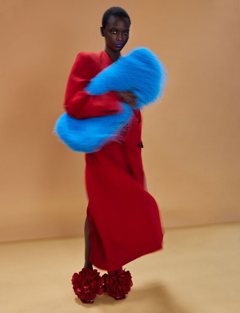 Model Oulimata wearing a long red coat and holding a bright cyan fuzzy stole against a warm beige studio backdrop.