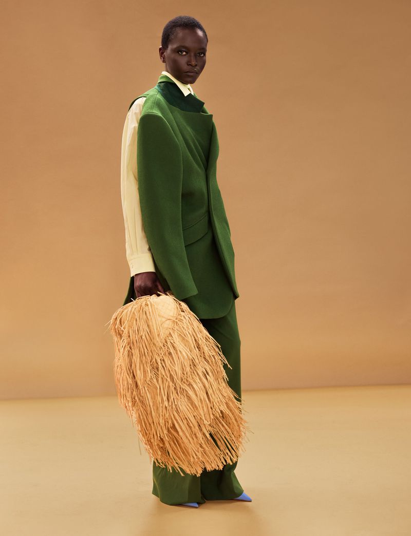 Model wearing a tailored green suit and pale yellow shirt holding a large straw fringe bag against a warm beige studio backdrop.