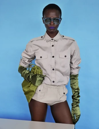 Model Oulimata wearing a beige utility shirt, olive green satin gloves, and blue tinted sunglasses against a solid blue backdrop.