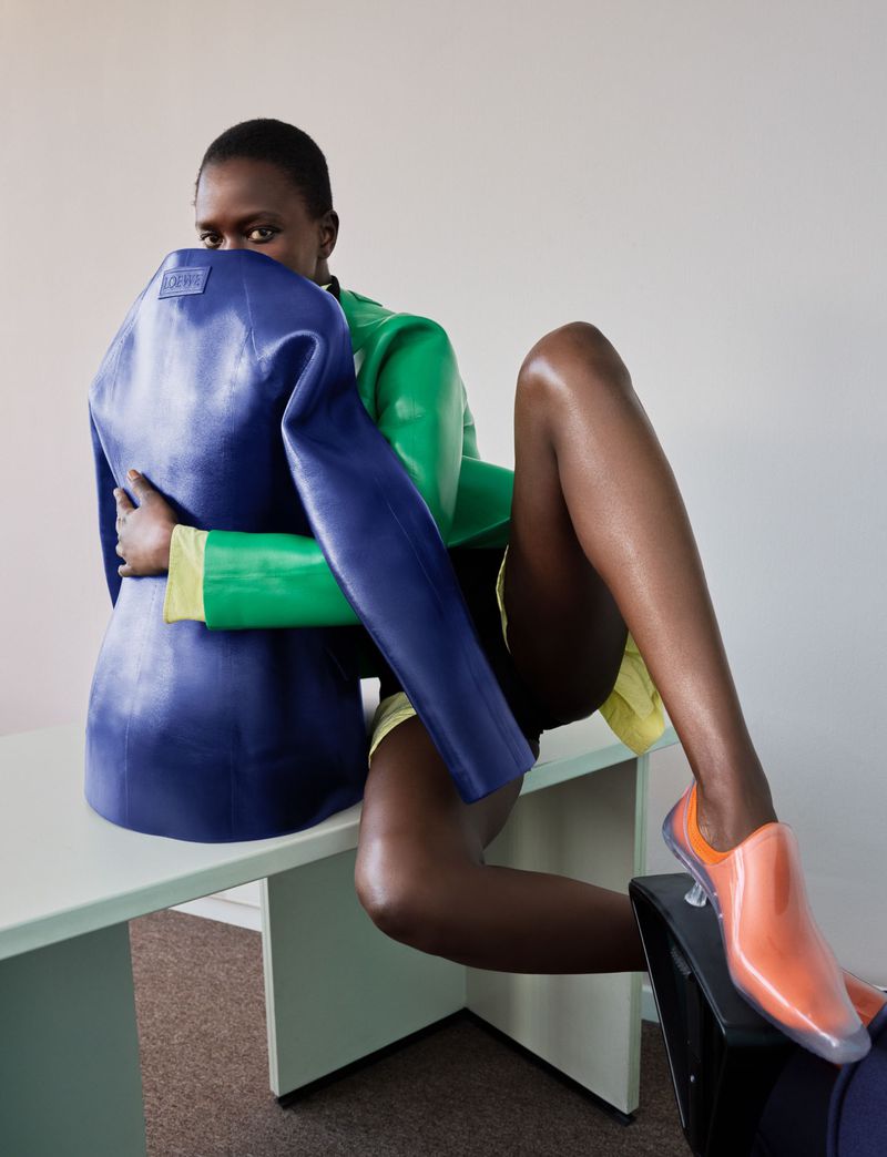 Model Oulimata holding a structured royal blue jacket while seated on a green desk in a green top and orange shoes.