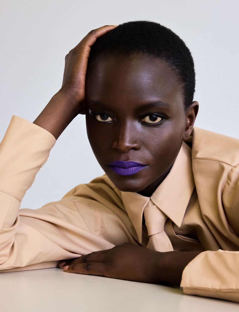 Close-up portrait of model Oulimata wearing bold purple lipstick, a beige collared shirt, and a matching necktie.
