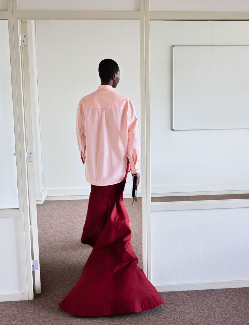 Model seen from behind wearing a pastel pink button-down shirt and a dramatic floor-length burgundy skirt in an office interior.