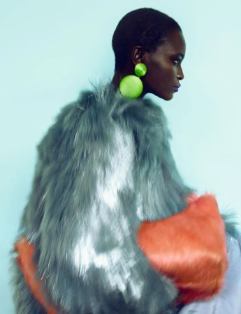 Model Oulimata in profile wearing a voluminous grey faux fur coat with bright green drop earrings against a light blue backdrop.