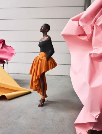 Model posing in a black off-shoulder top and sculptural asymmetric orange ruffled skirt with large pink and yellow paper props.