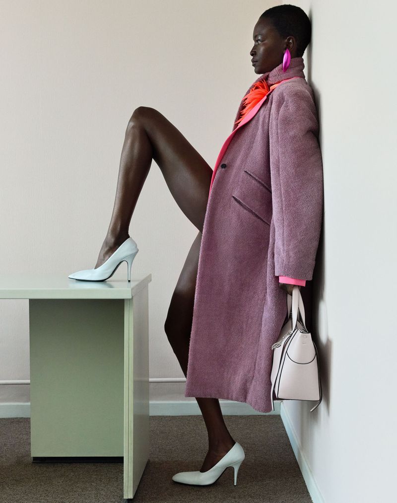 Model in mauve coat leaning against a white wall with one leg raised on a light green desk for Knack Weekend