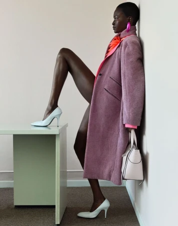 Model in mauve coat leaning against a white wall with one leg raised on a light green desk for Knack Weekend