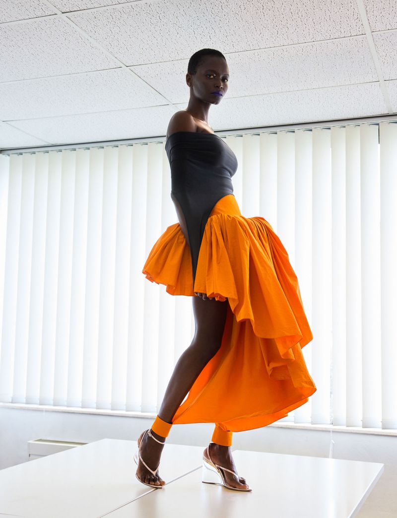 Fashion model in a black off-shoulder top and voluminous ruffled orange skirt standing on a table in front of vertical blinds.