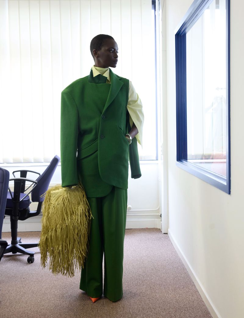 Model Oulimata standing in an office wearing an oversized green suit and holding a textured fringe handbag.