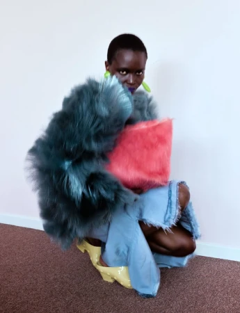 Model crouching against a plain wall wearing a voluminous teal faux fur coat, frayed denim, and holding a pink fuzzy bag.