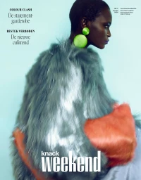 Model Oulimata wearing a grey shaggy fur coat with orange trim and bright green spherical earrings against a light blue backdrop.