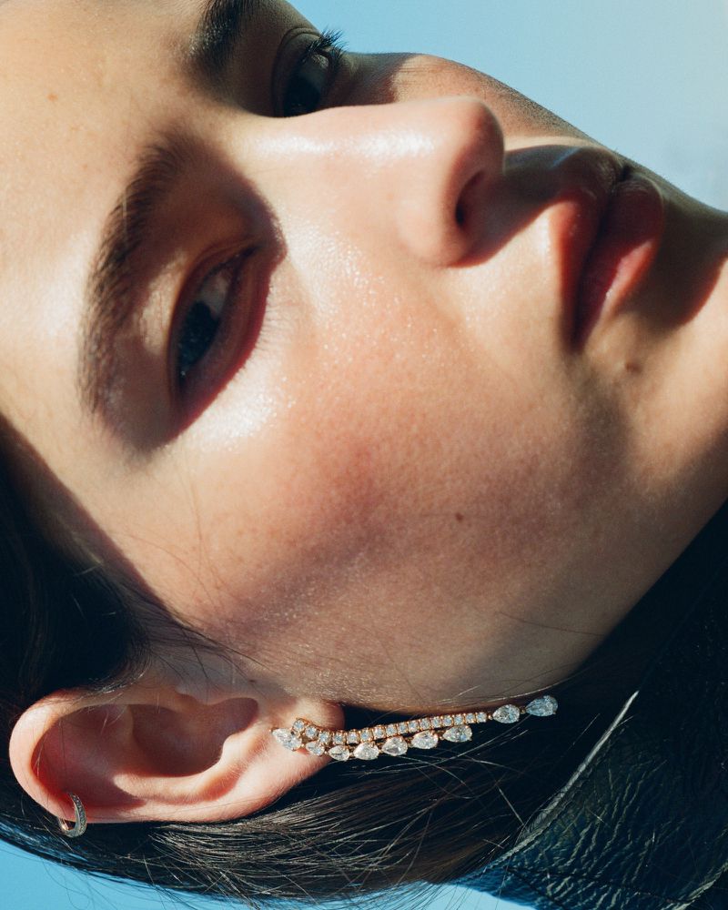 Close-up portrait of model Luiza Perote wearing a diamond ear climber earring in direct sunlight under a clear blue sky.