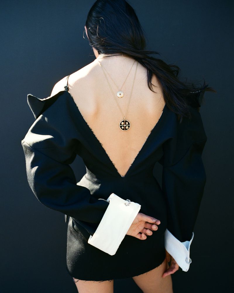 Model Luiza Perote viewed from behind in a black backless mini dress with wide white cuffs and gold medallion necklaces against a dark backdrop.