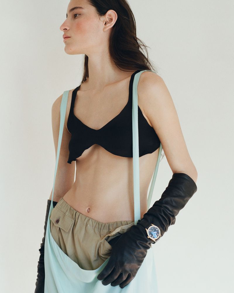 Model Luiza Perote wearing a black cropped top, long black leather gloves, and khaki pants against an off-white background.
