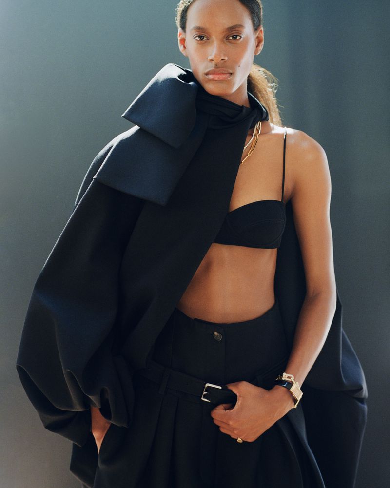 Model Luiza Perote wearing a sculptural draped black coat, black bra top, and tailored high-waisted trousers against a neutral backdrop.