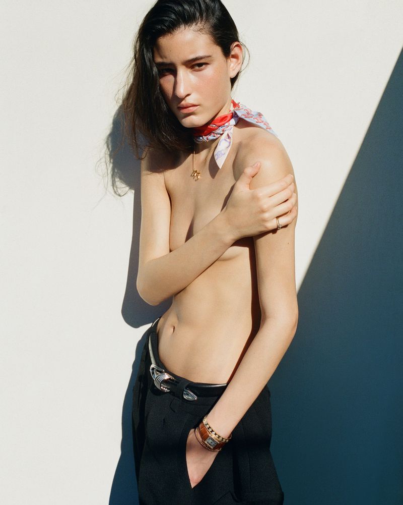 Model Luiza Perote posing topless in black trousers with a patterned neck scarf against a white wall with strong diagonal shadows.
