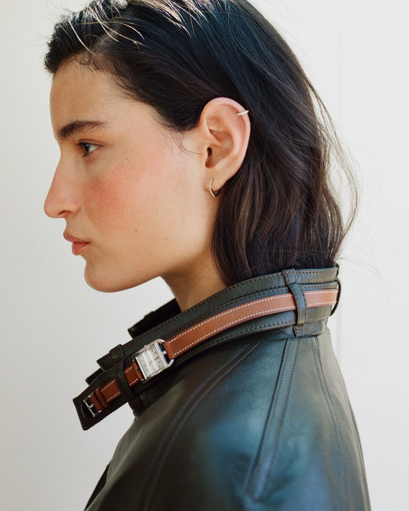 Side profile of model Luiza Perote wearing a dark leather jacket styled with a brown leather wristwatch around the collar.